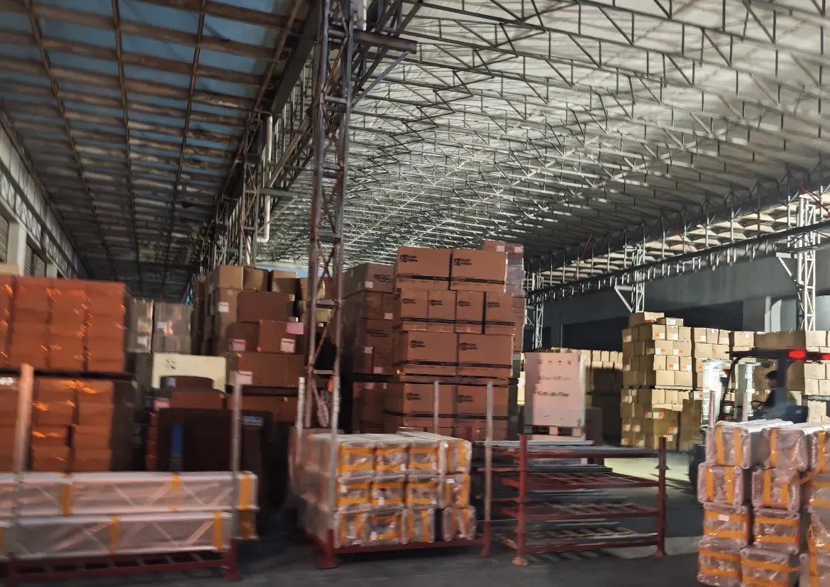 Stacked cartons and shrink-wrapped pallets under a covered warehouse bay, with a forklift working at the far end