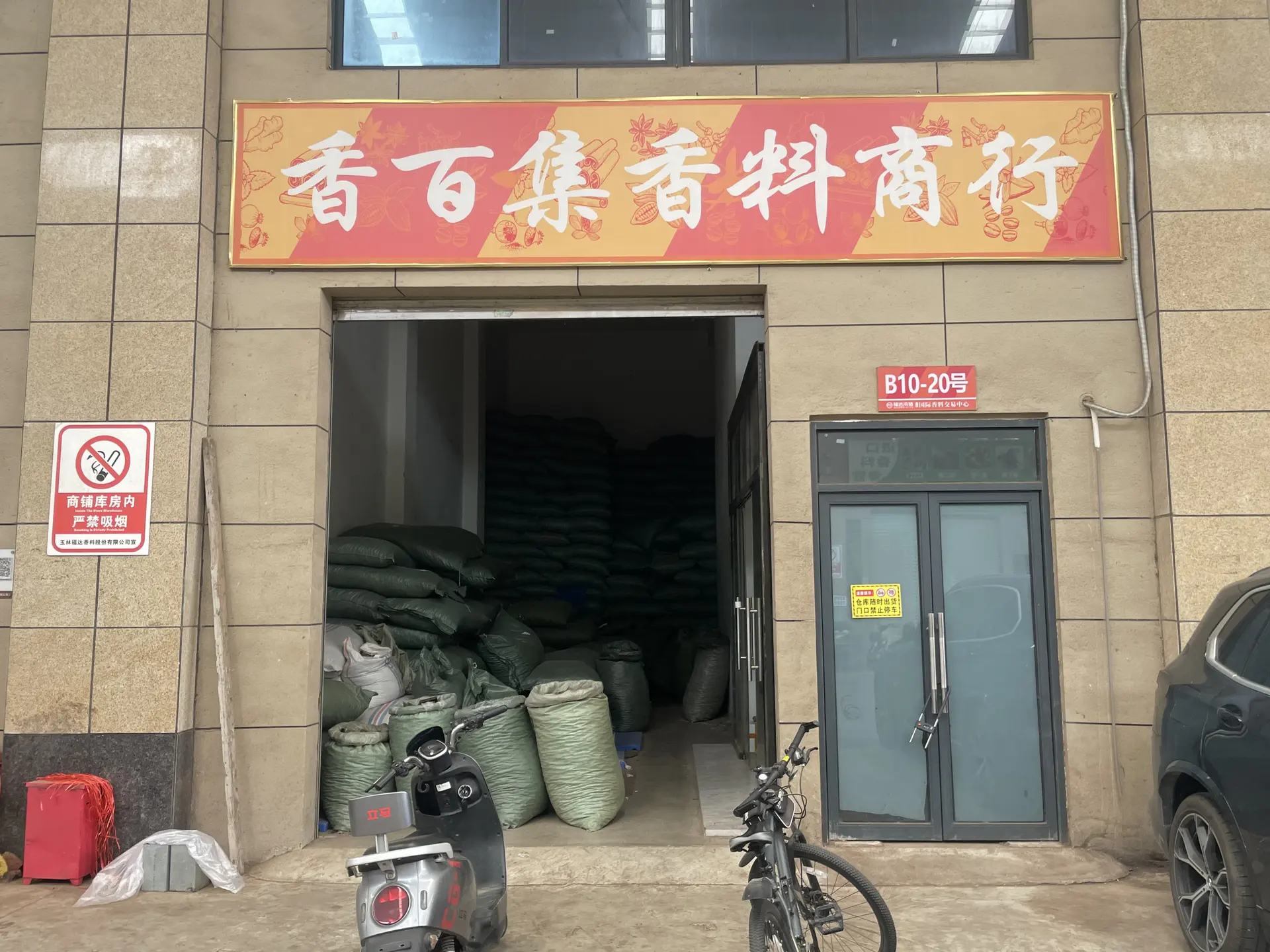 Fachada con el cartel de la tienda de especias Xiangbaiji sobre una puerta enrollable abierta, sacos de especias apilados dentro, el número de unidad B10-20 junto a la puerta